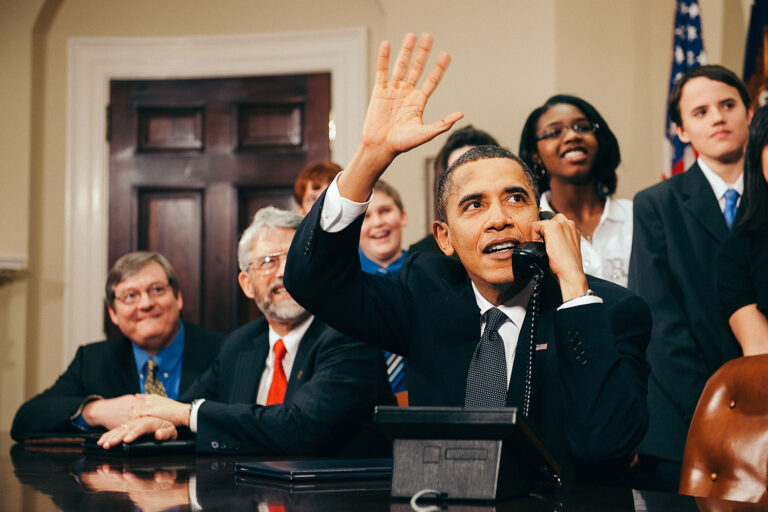 Barack Obama waving and talking on the phone surrounded by people