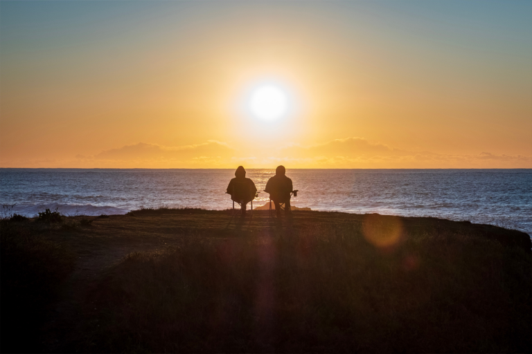 A couple sit in chairs on a cliff watching a beach sunset