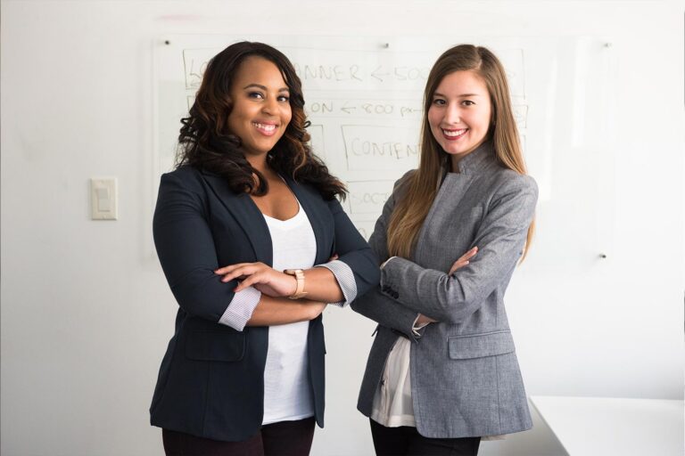 Two businesswomen stand with arms crossed in front of a whiteboard