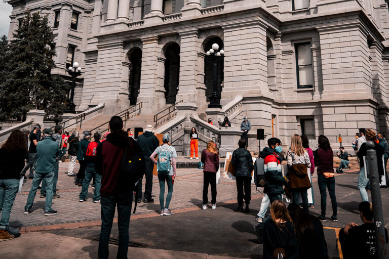 a crowd outside a government building listens to a speaker