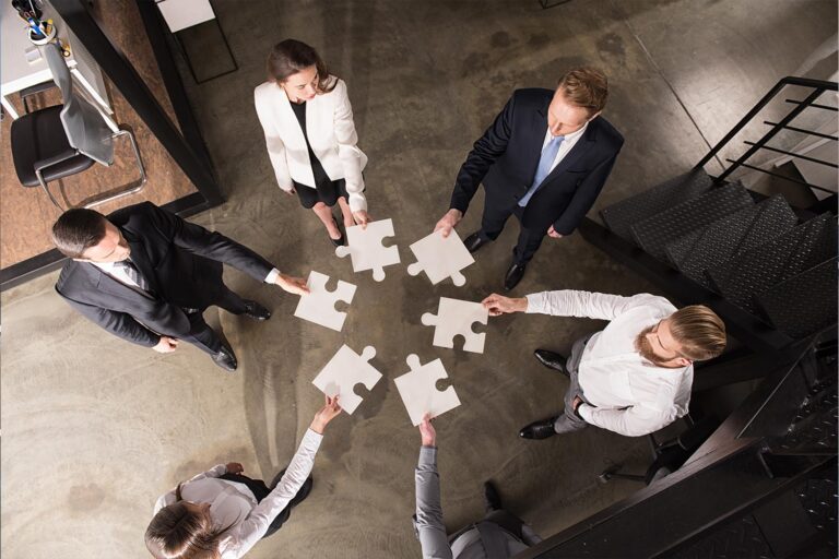 A group of businesspeople stand in a circle each holds out a puzzle piece