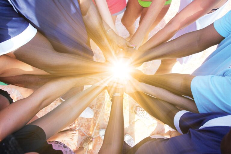 A group of people huddle with hands in the center, a light emanates from the middle