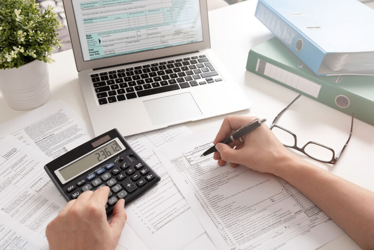 a desktop with hands writing and using an adding machine and a laptop and glasses also visible