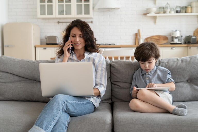 a woman working on a laptop and talking on the phone on a sofa next to a boy looking at a tablet