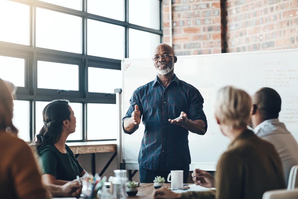 a person standing in front of a whiteboard speaking to a group of people seated around a table