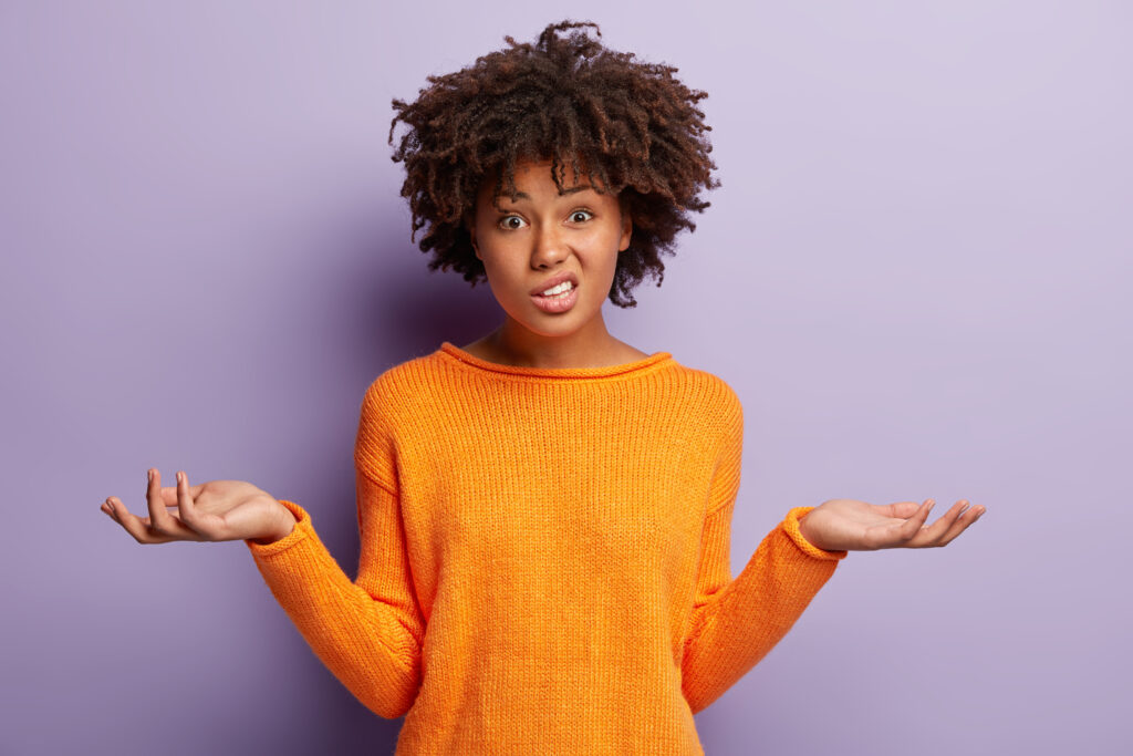 Isolated shot of confused beautiful woman with dark skin, curly haircut, spreads hands sideways, smirks face, feels doubt while makes choice, dressed in casual jumper, isolated over purple wall to illustrate nonprofit fundraising 